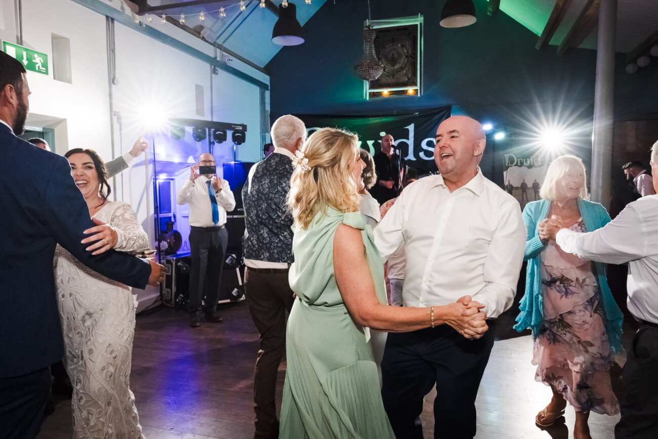 A Relaxed Summer Wedding at Village Barn, Tyrellspass | Amy & Eoghan 112 People are dancing in a brightly lit indoor venue, dressed in formal attire, with DJ equipment and decorations visible in the background.