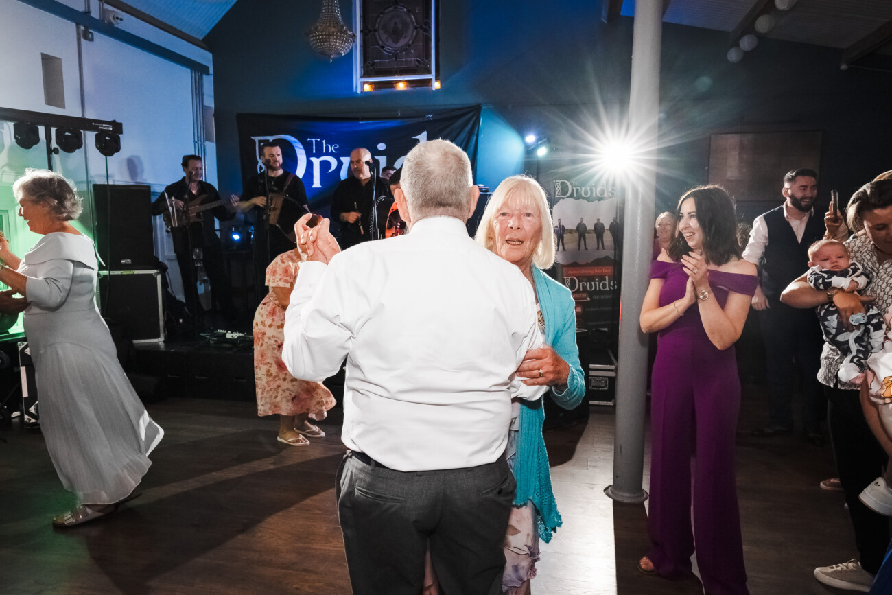 A Relaxed Summer Wedding at Village Barn, Tyrellspass | Amy & Eoghan 113 An elderly couple dances in the foreground at an indoor event whilst a live band plays on stage and guests watch and clap.