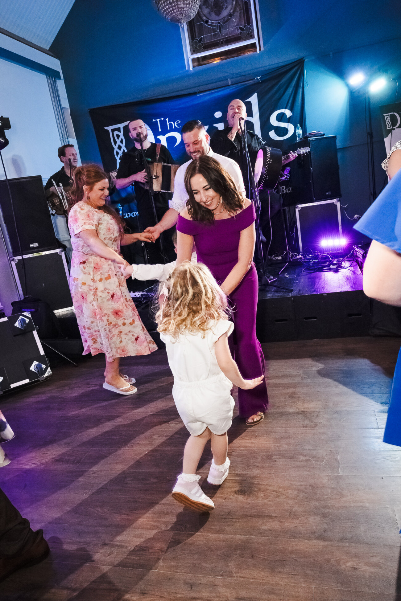 A Relaxed Summer Wedding at Village Barn, Tyrellspass | Amy & Eoghan 114 A woman in a purple dress dances with a young girl on a wooden floor whilst a band performs on stage in the background.