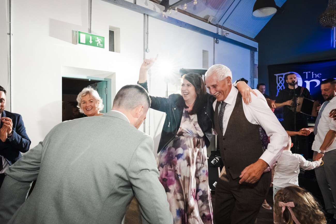 A Relaxed Summer Wedding at Village Barn, Tyrellspass | Amy & Eoghan 115 A group of people dance and smile at an indoor event, with adults in formal attire and a child in the foreground.