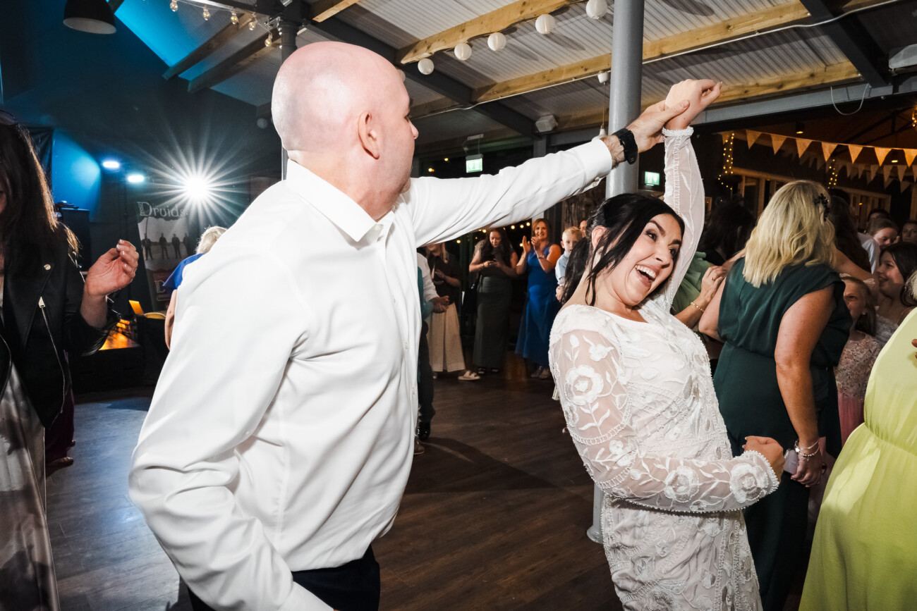 A Relaxed Summer Wedding at Village Barn, Tyrellspass | Amy & Eoghan 116 A man in a white shirt dances enthusiastically with a woman in a white dress at an indoor event, with other people in the background.