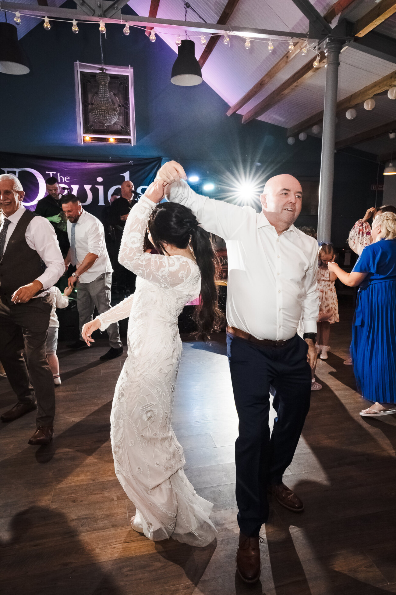 A Relaxed Summer Wedding at Village Barn, Tyrellspass | Amy & Eoghan 117 A man and woman, both dressed formally, dance together on a wooden floor at an indoor event whilst other guests watch and mingle in the background.