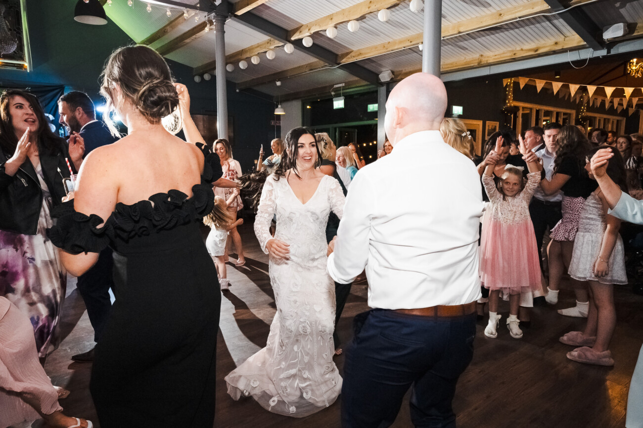 A Relaxed Summer Wedding at Village Barn, Tyrellspass | Amy & Eoghan 118 A bride in a white dress dances with guests at a wedding reception; people around them are clapping and smiling.