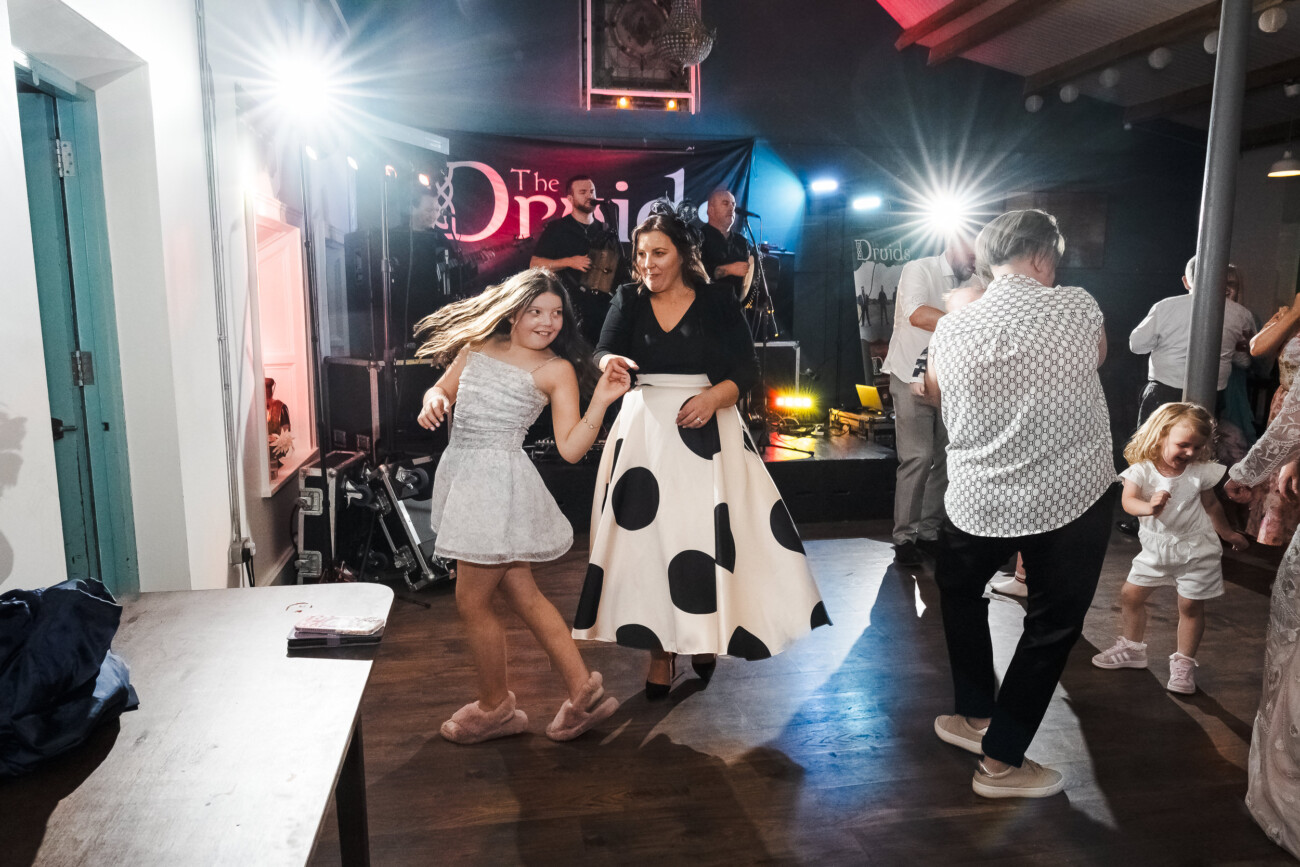 A Relaxed Summer Wedding at Village Barn, Tyrellspass | Amy & Eoghan 119 Two women dance in front of a live band on a stage at a party, with other people and children dancing nearby under bright lights.