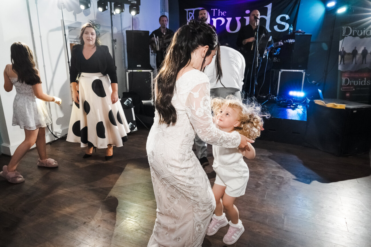 A Relaxed Summer Wedding at Village Barn, Tyrellspass | Amy & Eoghan 120 A woman in a white dress dances with a young girl at a party, whilst others dance nearby. A live band plays on stage in the background under a banner reading “Druids”.