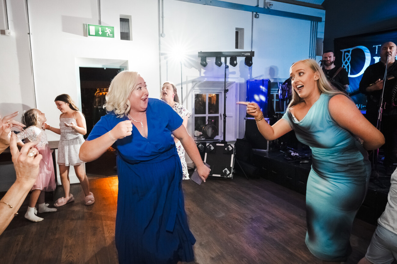 A Relaxed Summer Wedding at Village Barn, Tyrellspass | Amy & Eoghan 121 Two women enthusiastically dance and point at each other on a wooden floor, while other guests watch and smile in a brightly lit party setting.