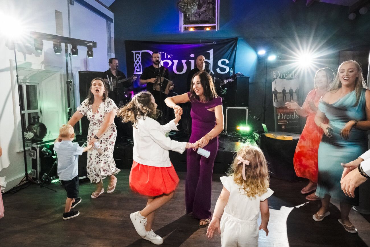 A Relaxed Summer Wedding at Village Barn, Tyrellspass | Amy & Eoghan 122 Adults and children dance together at an indoor event with a live band called “The Druids” performing on stage in the background.
