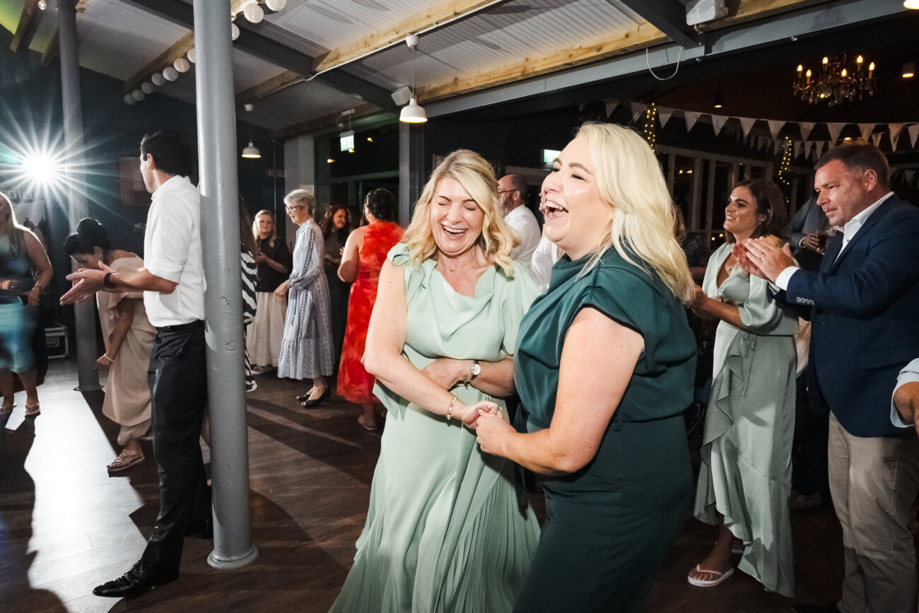 A Relaxed Summer Wedding at Village Barn, Tyrellspass | Amy & Eoghan 123 Two women in green dresses laugh and dance together at a lively indoor event, surrounded by other people who are applauding and enjoying the celebration.