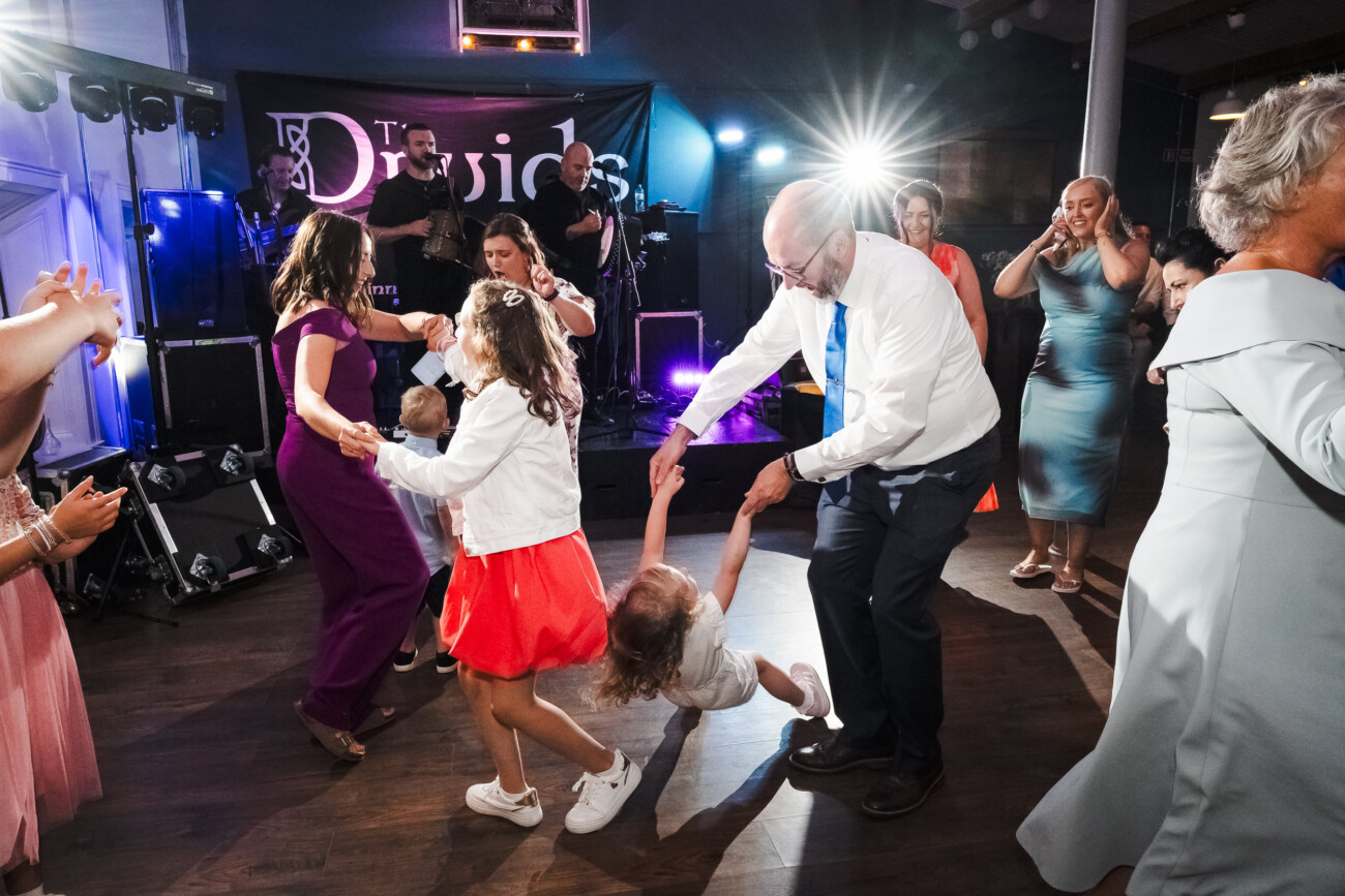 A Relaxed Summer Wedding at Village Barn, Tyrellspass | Amy & Eoghan 124 Adults and children dance energetically at an indoor event, with a live band performing in the background under bright lights.