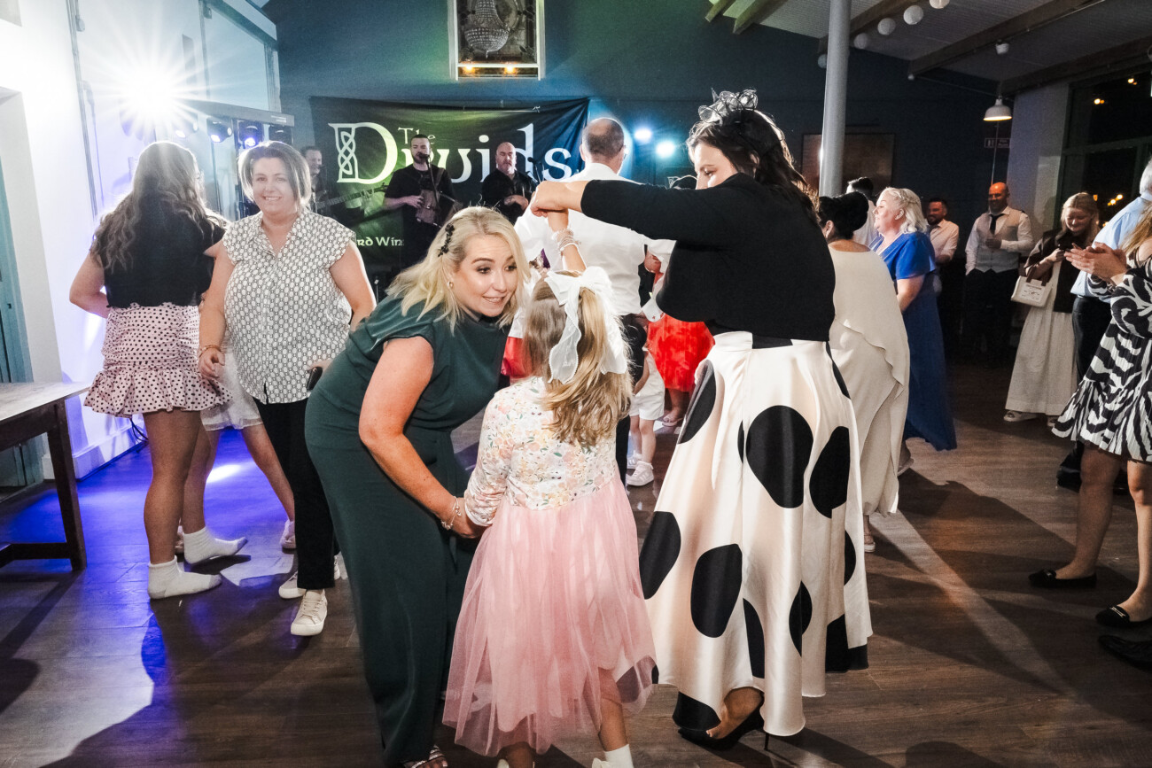A Relaxed Summer Wedding at Village Barn, Tyrellspass | Amy & Eoghan 125 Adults and children gather on a dance floor at a party, some talking and others dancing, with a green “Dividuals” sign visible in the background.