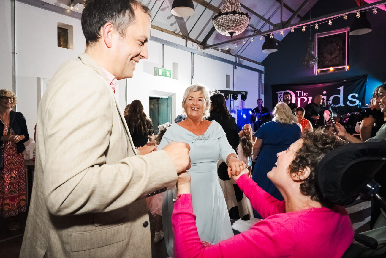 A Relaxed Summer Wedding at Village Barn, Tyrellspass | Amy & Eoghan 128 Three people hold hands and dance at an indoor event with a live band performing in the background. Other guests watch and mingle in the decorated venue.