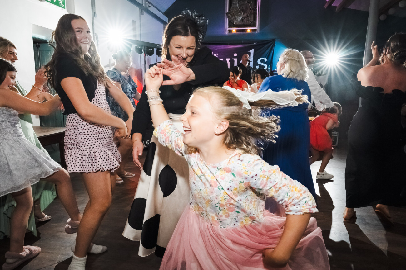 A Relaxed Summer Wedding at Village Barn, Tyrellspass | Amy & Eoghan 129 Children and adults dance energetically at an indoor party, illuminated by bright lights, with joyful expressions and colourful clothing visible throughout the lively crowd.