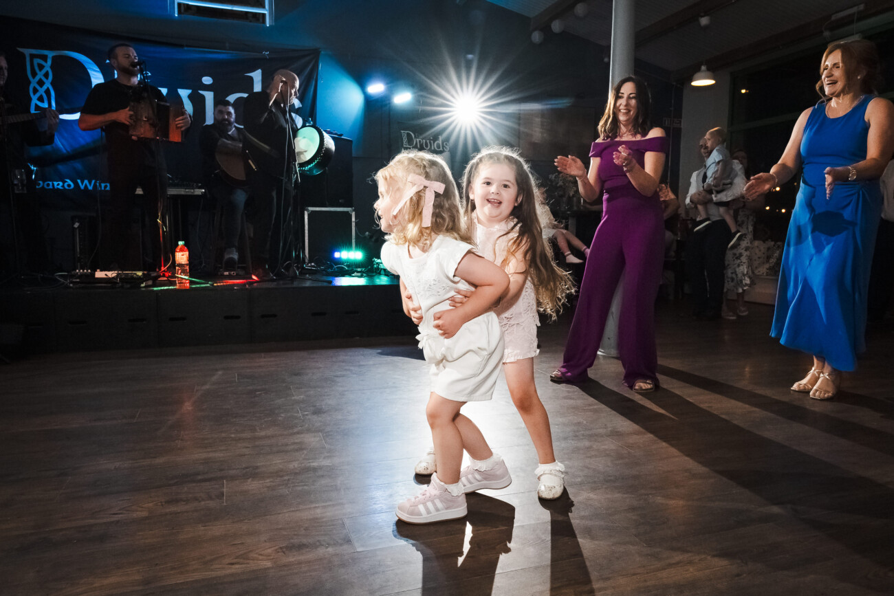 A Relaxed Summer Wedding at Village Barn, Tyrellspass | Amy & Eoghan 130 Two young girls smile and hug whilst dancing on a wooden floor at an indoor event, with a live band and adults clapping in the background.