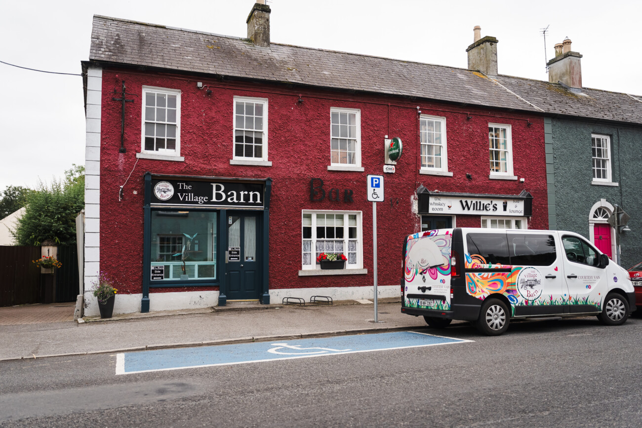 A Relaxed Summer Wedding at Village Barn, Tyrellspass | Amy & Eoghan 1 Red brick building with “The Village Barn” bar, “Willie’s” signage, and a colourful van parked in a blue parking space on the street in front.