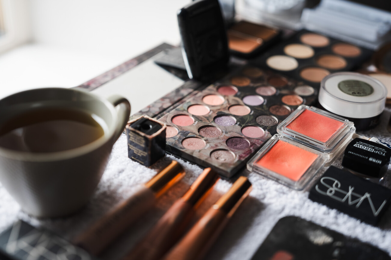 A Relaxed Summer Wedding at Village Barn, Tyrellspass | Amy & Eoghan 2 A close-up of assorted make-up products, including eyeshadow palettes, blusher, brushes, and a cup of tea on a white towel.