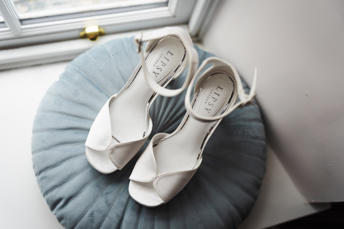 A Relaxed Summer Wedding at Village Barn, Tyrellspass | Amy & Eoghan 3 A pair of white open-toe sandals with ankle straps rests on a round, pleated blue cushion by a window.