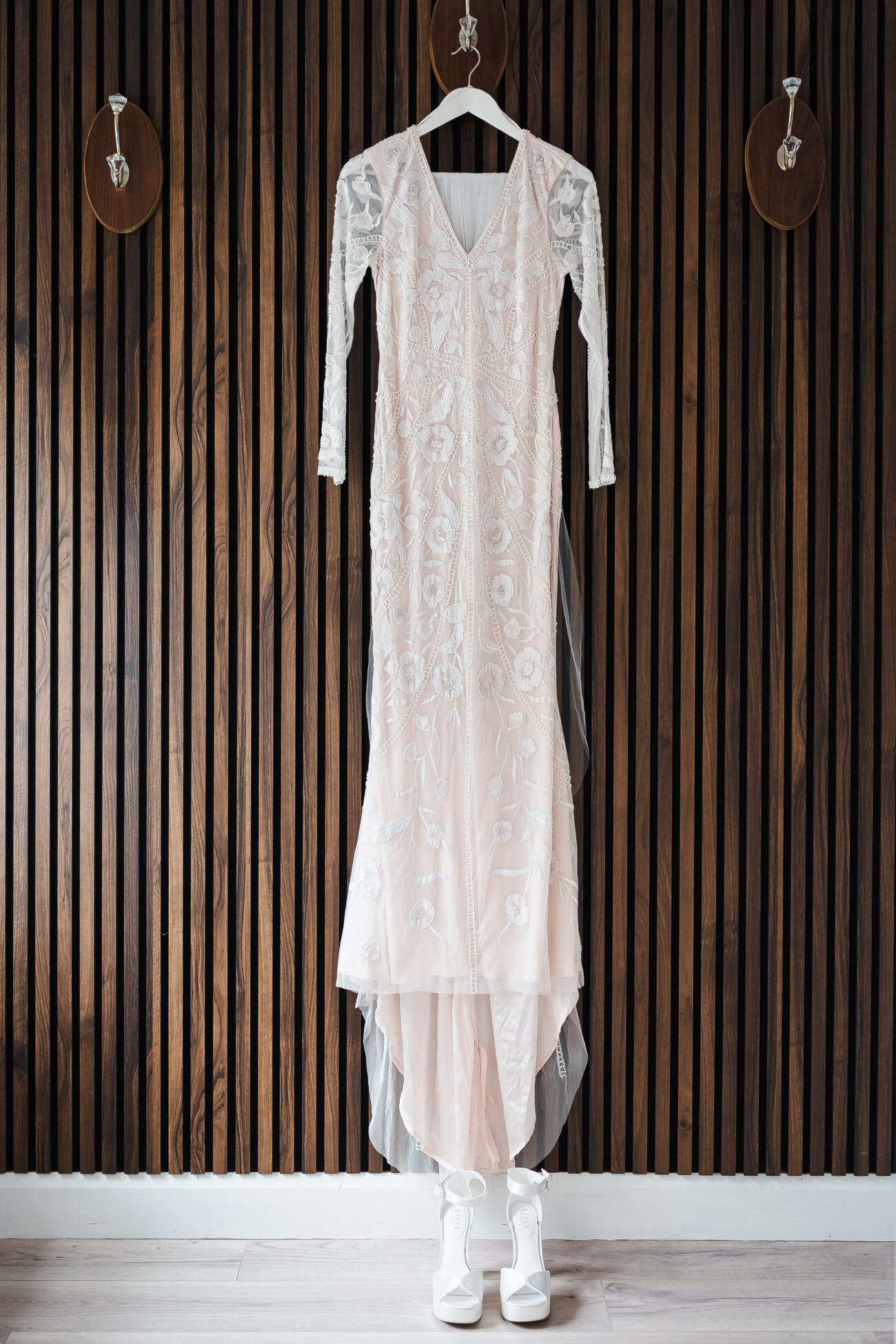 A Relaxed Summer Wedding at Village Barn, Tyrellspass | Amy & Eoghan 4 A long-sleeved, embroidered white dress hangs on a wooden wall above a pair of white boots, with three mounted hooks spaced evenly on the wall.