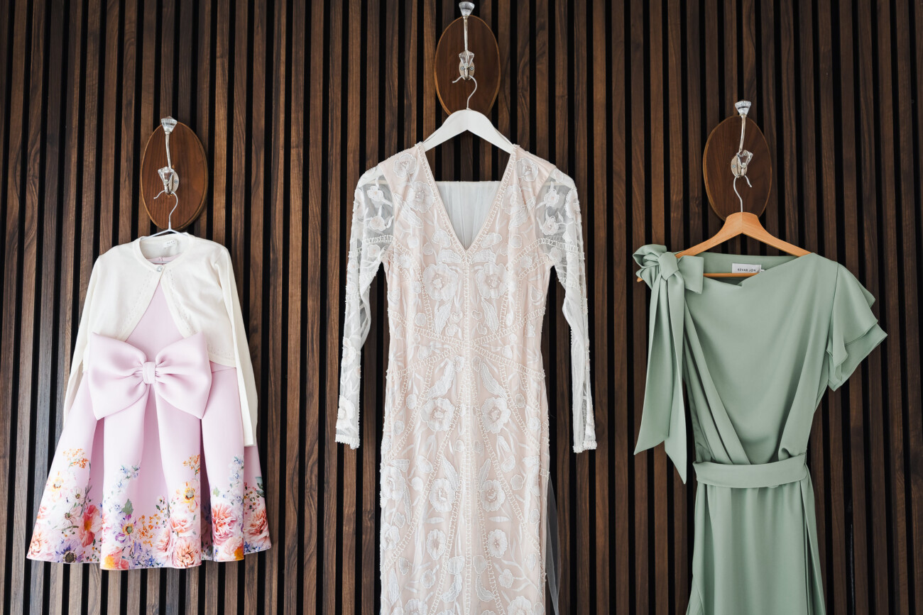 A Relaxed Summer Wedding at Village Barn, Tyrellspass | Amy & Eoghan 5 Three dresses on hangers against a wooden wall: a pink floral girls’ dress with a bow, a white lace long-sleeve dress, and a sage green dress with a side bow.