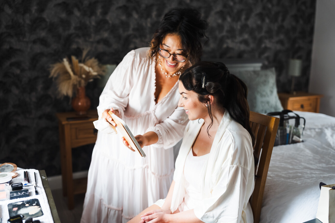 A Relaxed Summer Wedding at Village Barn, Tyrellspass | Amy & Eoghan 6 Two women in light-coloured robes smile whilst looking at a tablet in a well-lit bedroom. A dressing table with various products is visible in the foreground.