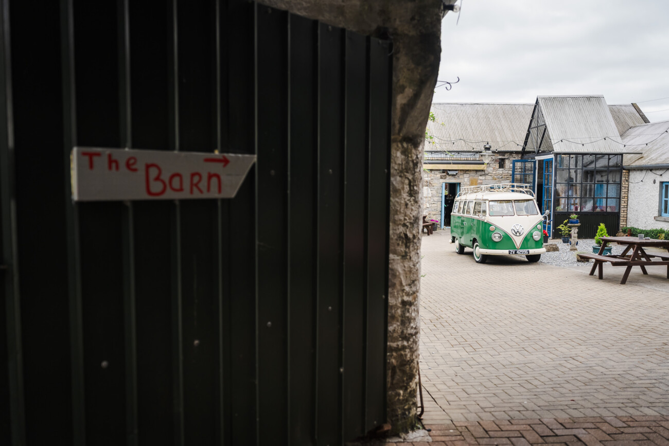 A Relaxed Summer Wedding at Village Barn, Tyrellspass | Amy & Eoghan 7 A sign reading “The Barn” points to a courtyard with a green and white vintage van parked near stone buildings and picnic tables.