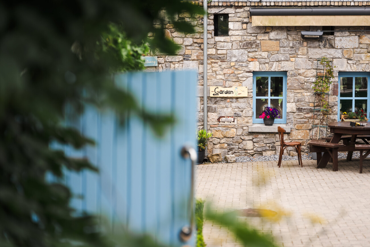 A Relaxed Summer Wedding at Village Barn, Tyrellspass | Amy & Eoghan 8 A stone building with blue-framed windows, a wooden bench and table, potted flowers, and a “Garden” sign is seen beyond a partially open blue gate.