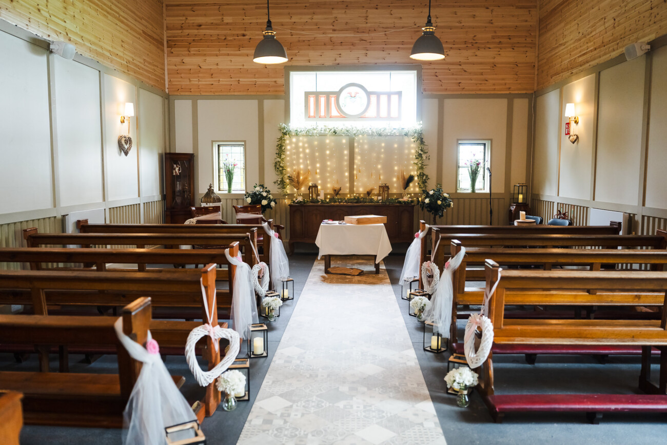 A Relaxed Summer Wedding at Village Barn, Tyrellspass | Amy & Eoghan 10 A small chapel is decorated for a wedding, with wooden pews, heart-shaped ornaments, flowers, and white fabric. The altar is adorned with lights and floral arrangements.