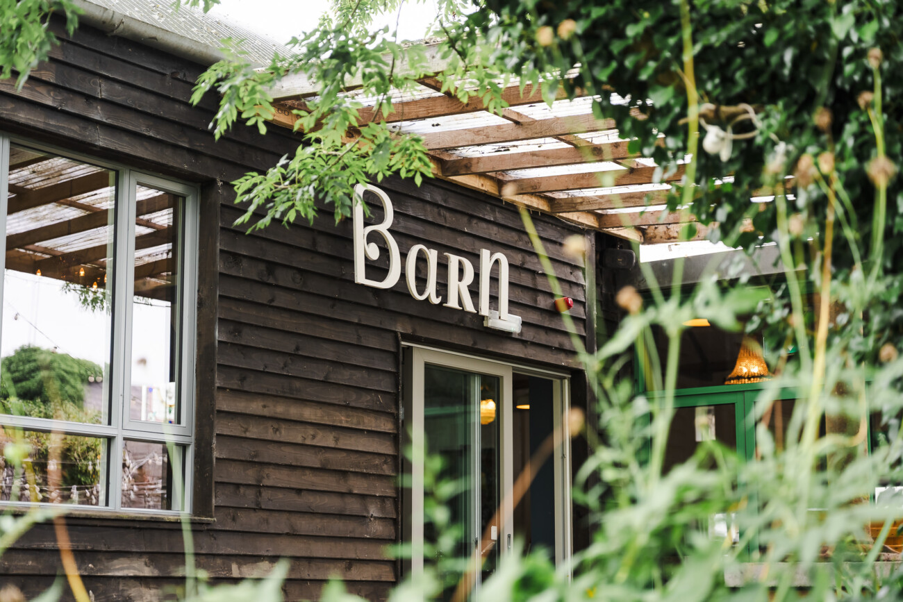 A Relaxed Summer Wedding at Village Barn, Tyrellspass | Amy & Eoghan 11 Wooden building with the word “Barn” in large letters on the side, partially obscured by green plants in the foreground.