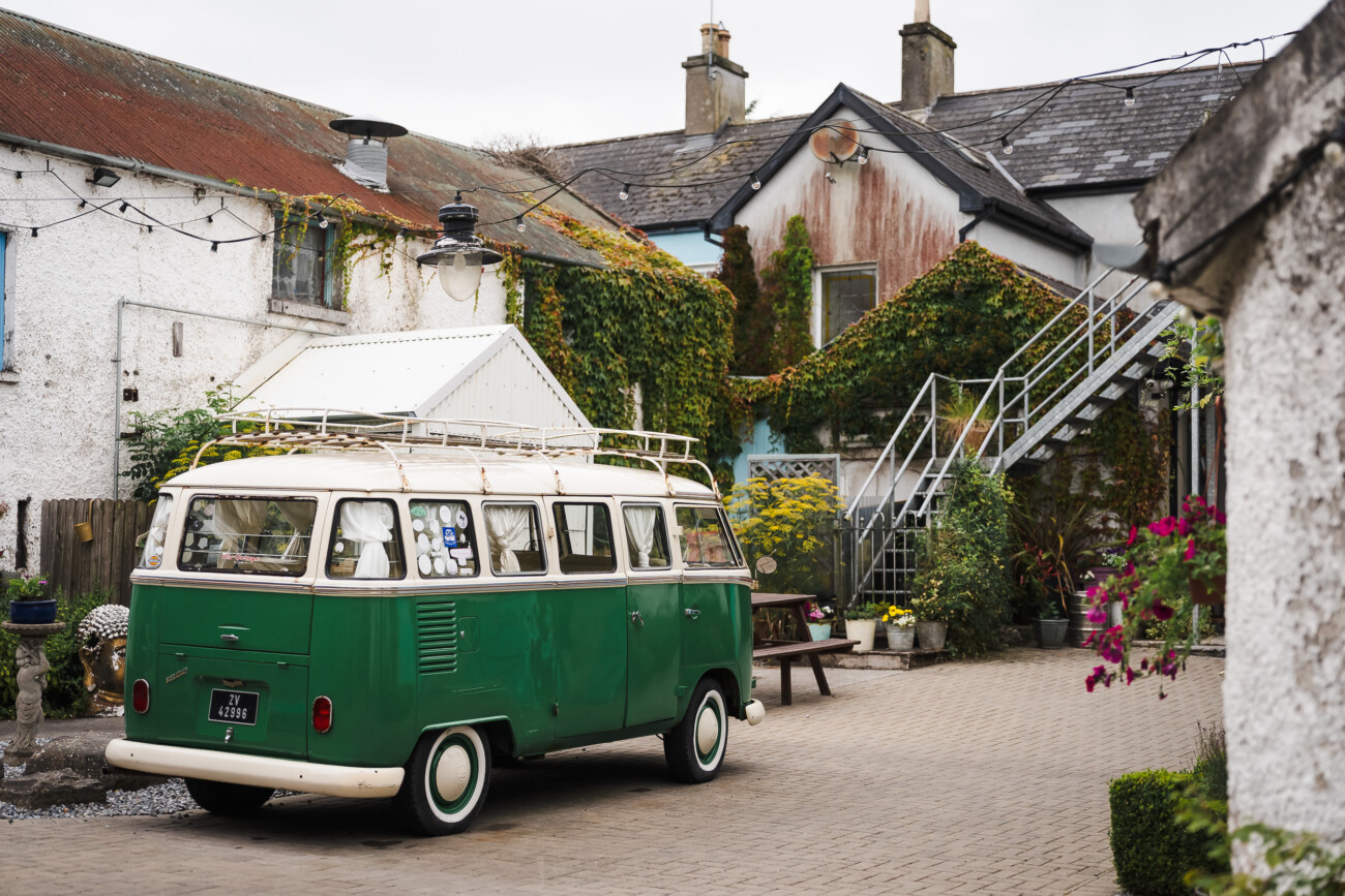 A Relaxed Summer Wedding at Village Barn, Tyrellspass | Amy & Eoghan 12 A green and white vintage van is parked in a cobbled courtyard surrounded by ivy-covered white buildings with outdoor stairs and plants.