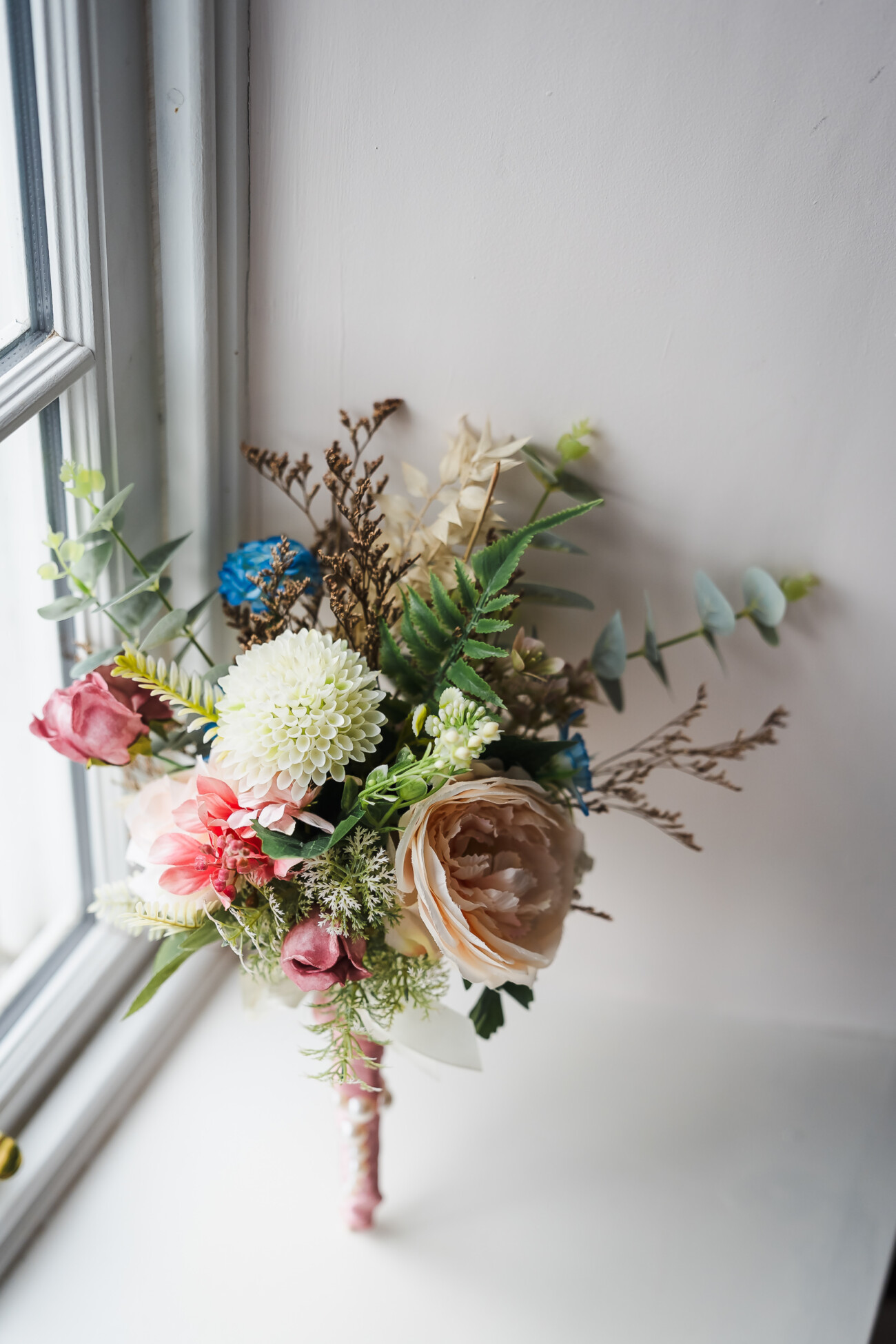 A Relaxed Summer Wedding at Village Barn, Tyrellspass | Amy & Eoghan 13 A bouquet of assorted flowers, including roses and chrysanthemums, rests on a white windowsill by a pale wall, with natural light coming from the window.