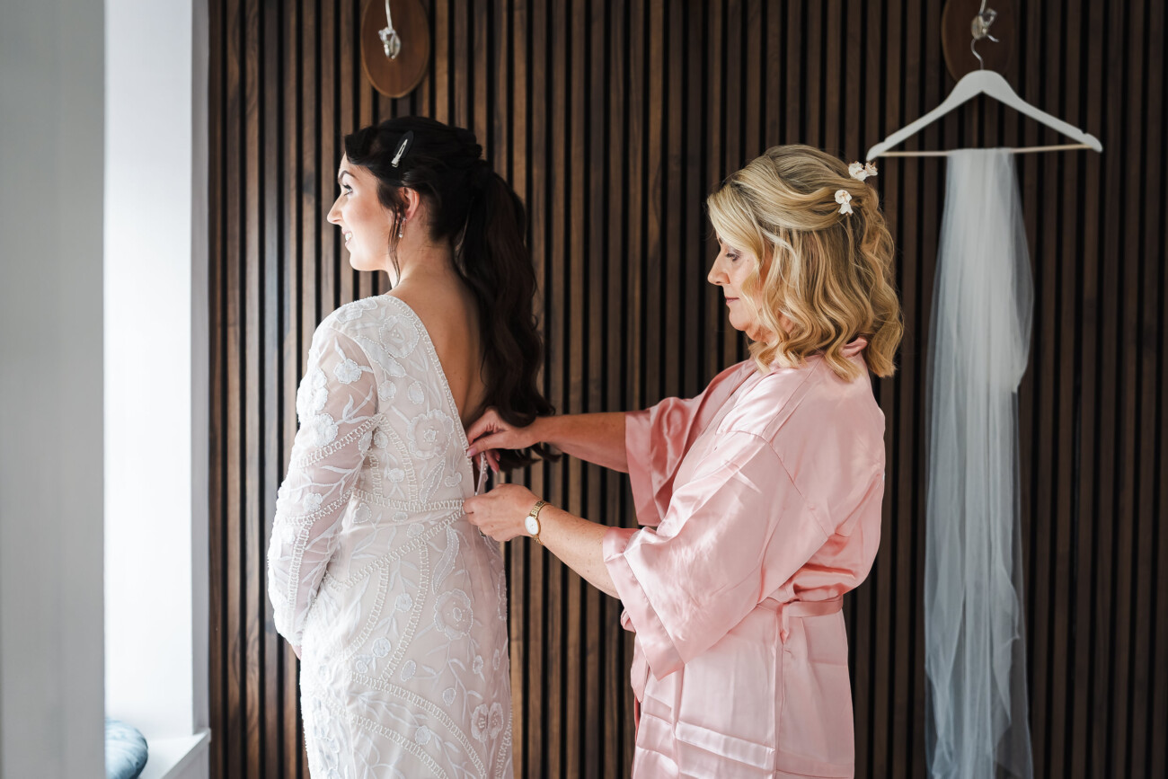 A Relaxed Summer Wedding at Village Barn, Tyrellspass | Amy & Eoghan 14 Woman in a pink dressing gown fastens the back of a white dress for another woman standing by a wooden wall, with a veil hanging nearby.