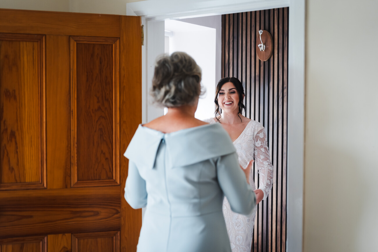 A Relaxed Summer Wedding at Village Barn, Tyrellspass | Amy & Eoghan 15 A woman in a white dress smiles as she faces another woman in a light blue dress standing in a doorway.