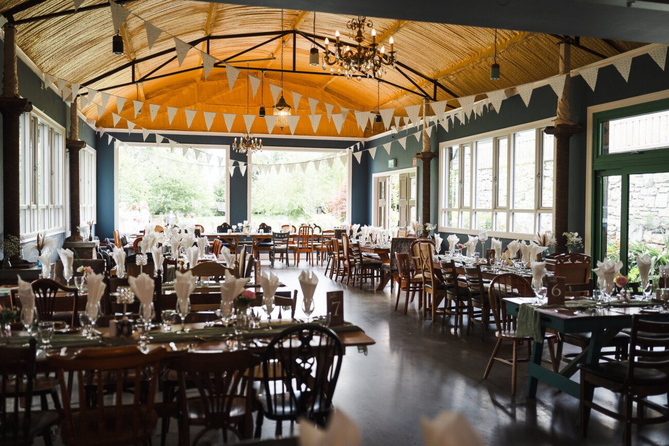 A Relaxed Summer Wedding at Village Barn, Tyrellspass | Amy & Eoghan 16 Large event hall with wooden ceiling, decorated with white bunting. Tables are set with glassware, plates, and folded serviettes, ready for a formal gathering. Natural light fills the room.