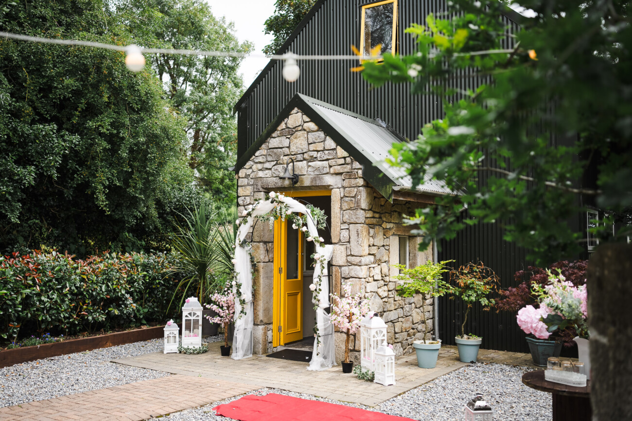 A Relaxed Summer Wedding at Village Barn, Tyrellspass | Amy & Eoghan 17 Stone building with a yellow door, decorated white arch, potted plants, lanterns, and a red carpet, set in a garden with trees and bushes visible in the background.