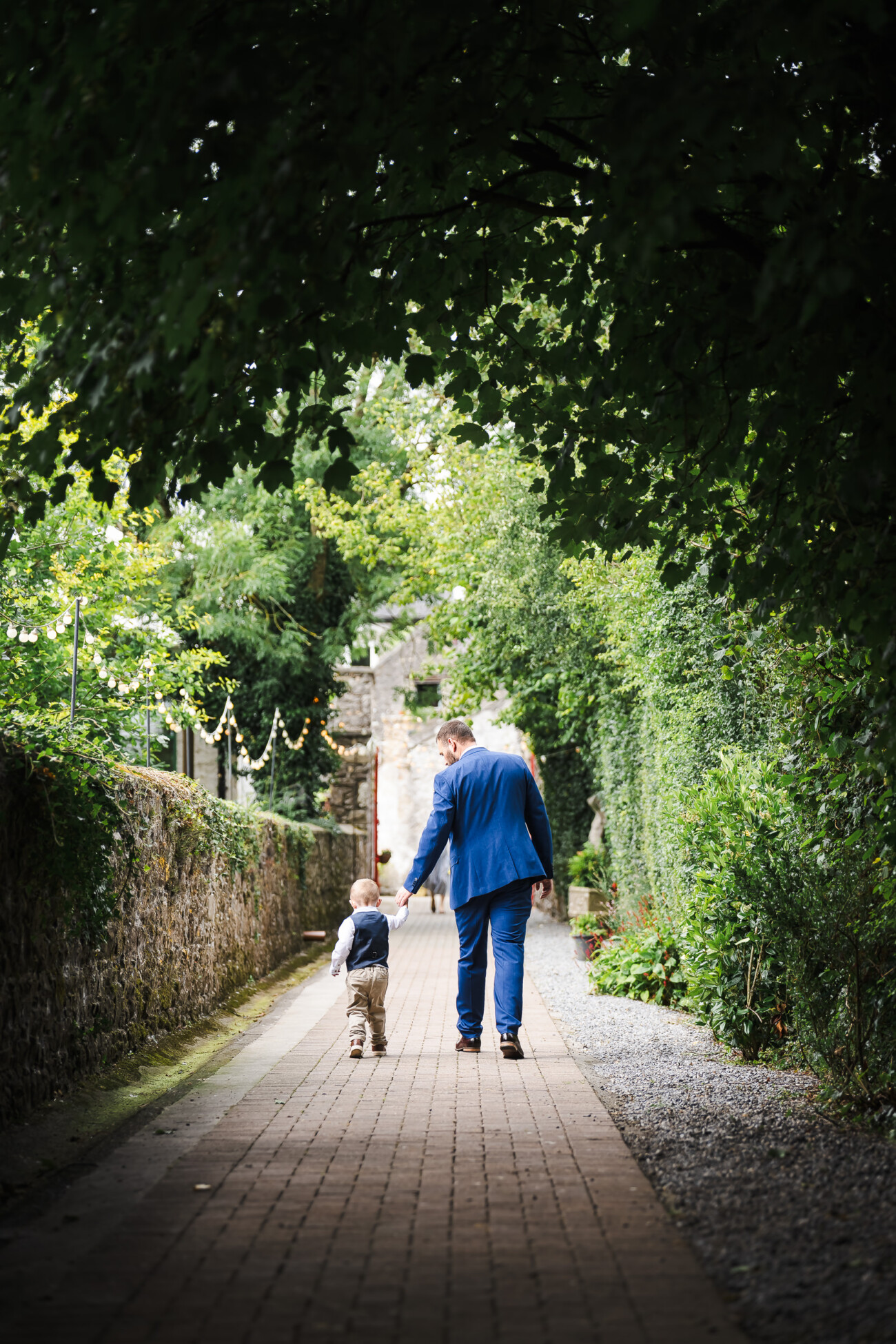 A Relaxed Summer Wedding at Village Barn, Tyrellspass | Amy & Eoghan 18 Man in a blue suit holds hands with a small child as they walk down a brick path lined with greenery, facing away from the camera.