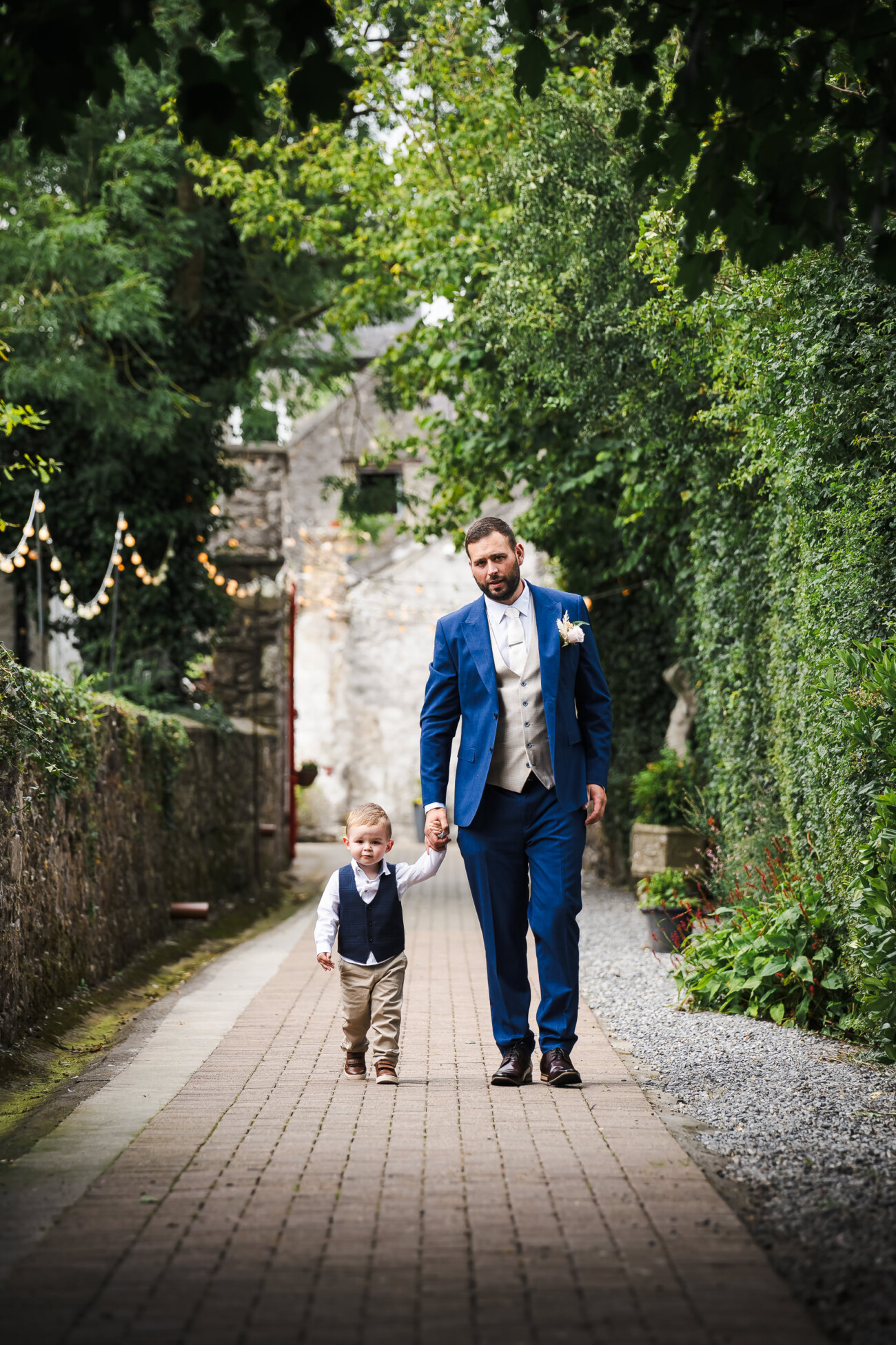 A Relaxed Summer Wedding at Village Barn, Tyrellspass | Amy & Eoghan 19 A man in a blue suit walks hand in hand with a young boy in formal attire down a brick path surrounded by greenery.