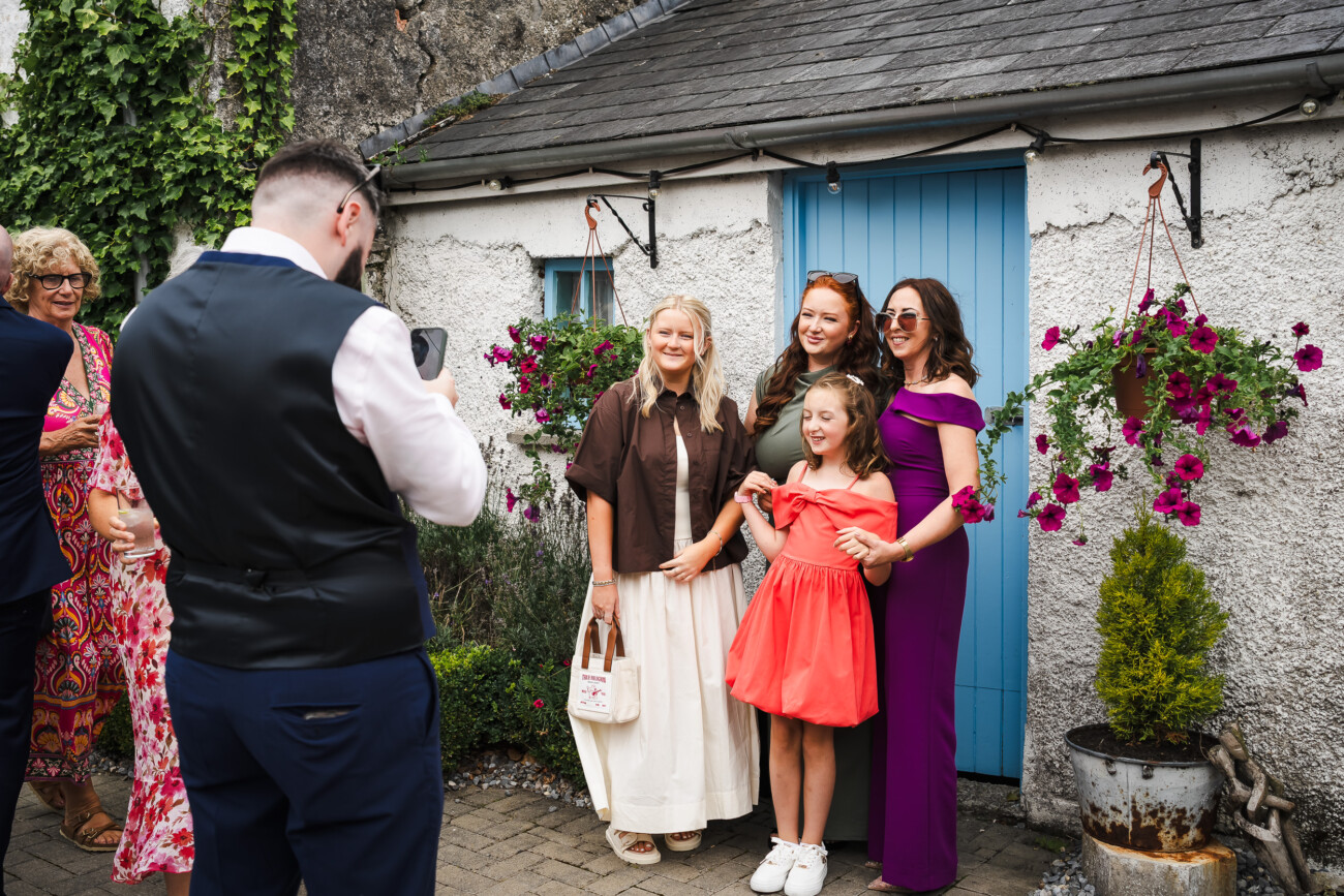 A Relaxed Summer Wedding at Village Barn, Tyrellspass | Amy & Eoghan 21 A man takes a photo of four people posing and smiling in front of a blue door, with hanging flower baskets and other people in the background.