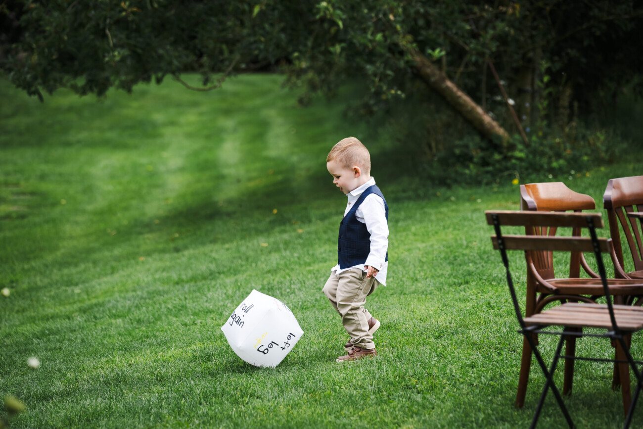 A Relaxed Summer Wedding at Village Barn, Tyrellspass | Amy & Eoghan 23 A young boy in smart clothes stands on grass next to a large white balloon, with empty wooden chairs and trees in the background.