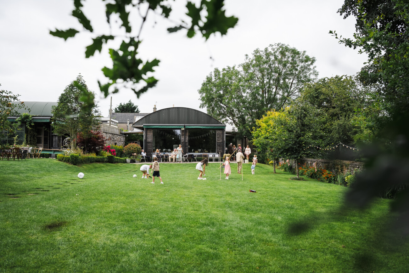 A Relaxed Summer Wedding at Village Barn, Tyrellspass | Amy & Eoghan 24 Children play on a large, grassy lawn in front of a modern barn-style building, with adults seated nearby and trees and garden beds surrounding the area.