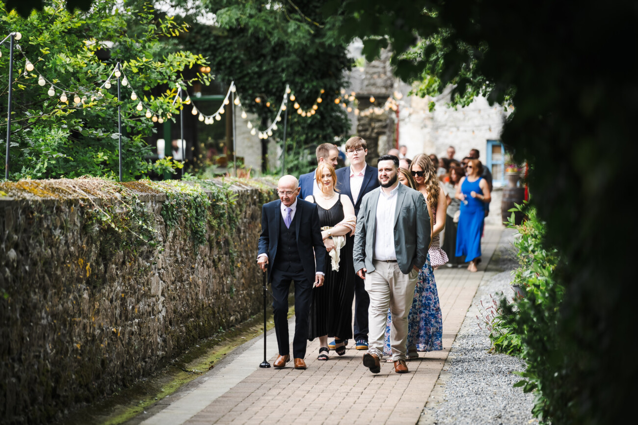 A Relaxed Summer Wedding at Village Barn, Tyrellspass | Amy & Eoghan 25 A group of people dressed formally walk along an outdoor path lined with stone walls and fairy lights during what appears to be a special event.