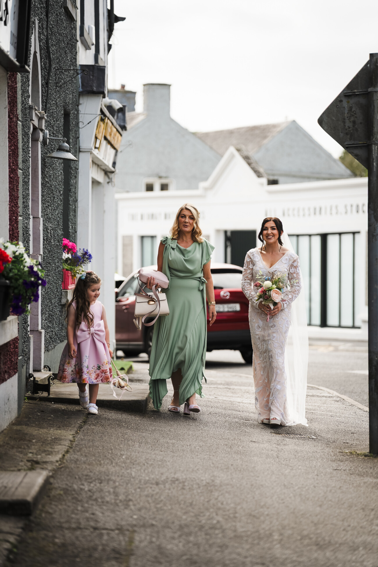 A Relaxed Summer Wedding at Village Barn, Tyrellspass | Amy & Eoghan 26 A bride in a white dress walks outdoors with a woman in green and a young girl in a pink dress, approaching a building on a street.