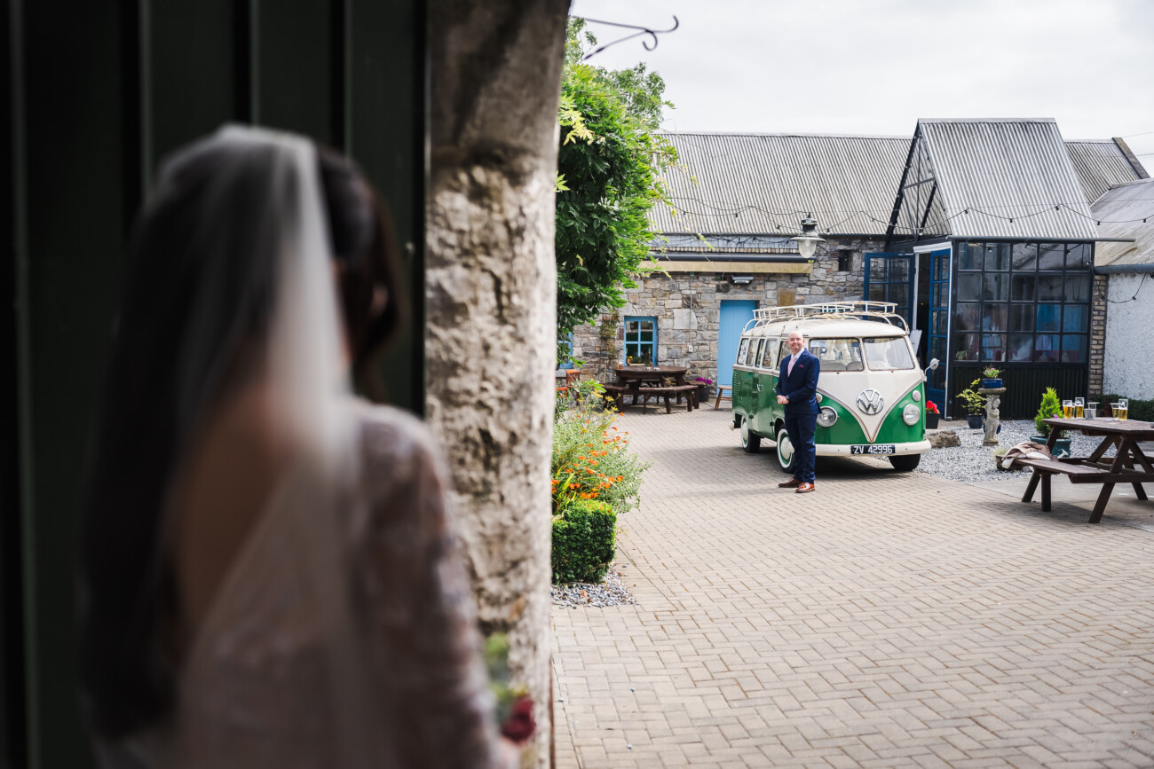 A Relaxed Summer Wedding at Village Barn, Tyrellspass | Amy & Eoghan 27 A bride stands in the foreground while a man in a suit stands near a vintage green and white van outside rustic buildings.