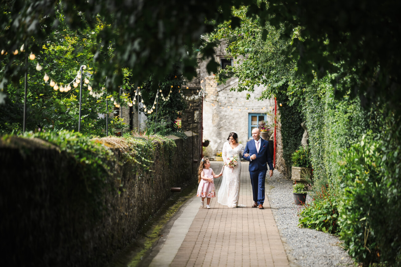A Relaxed Summer Wedding at Village Barn, Tyrellspass | Amy & Eoghan 28 A bride walks on a garden path accompanied by a man in a suit and a young girl, surrounded by greenery and fairy lights.