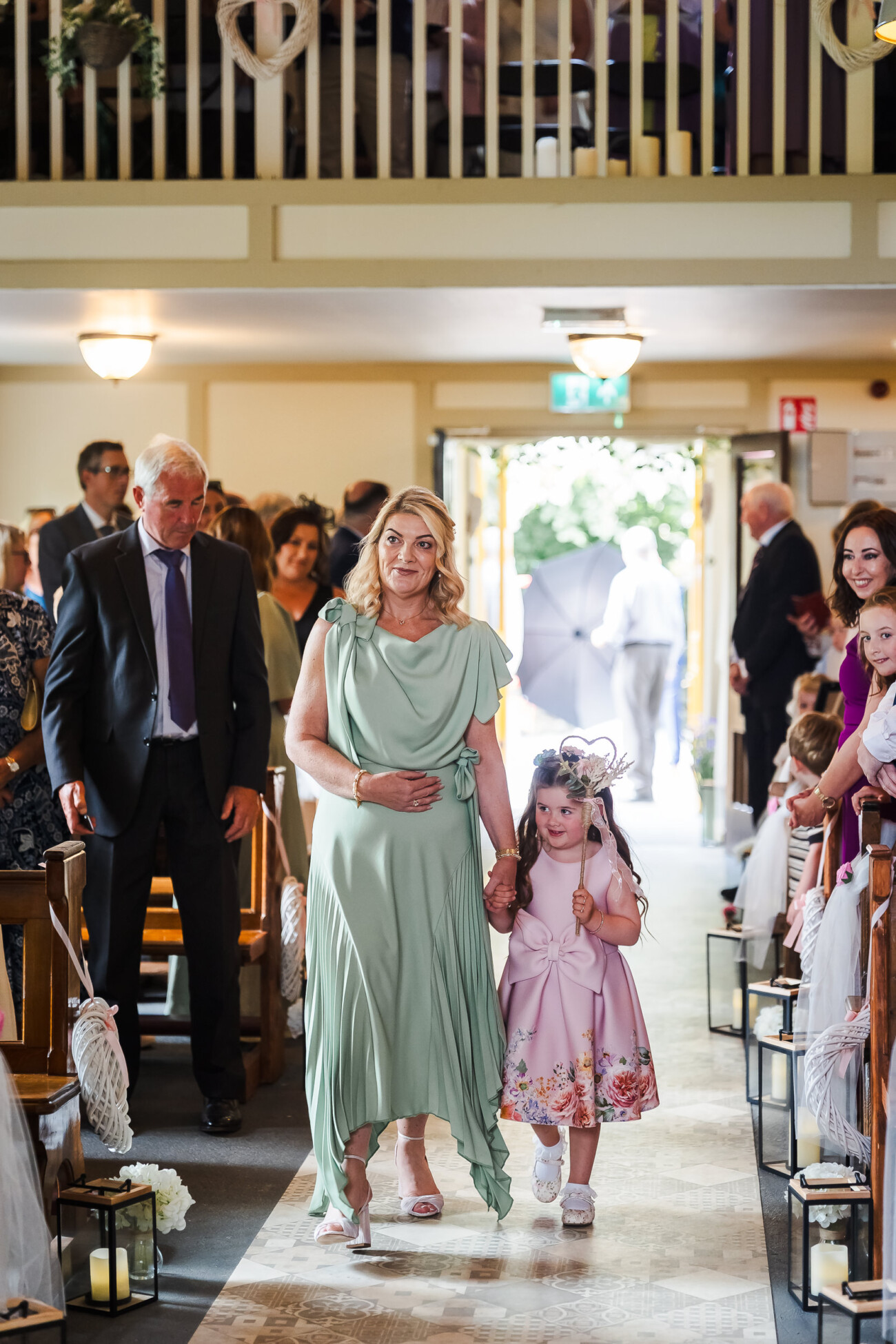 A Relaxed Summer Wedding at Village Barn, Tyrellspass | Amy & Eoghan 29 A woman in a light green dress walks down the aisle holding hands with a young girl in a pink dress at a wedding ceremony, with seated guests watching.