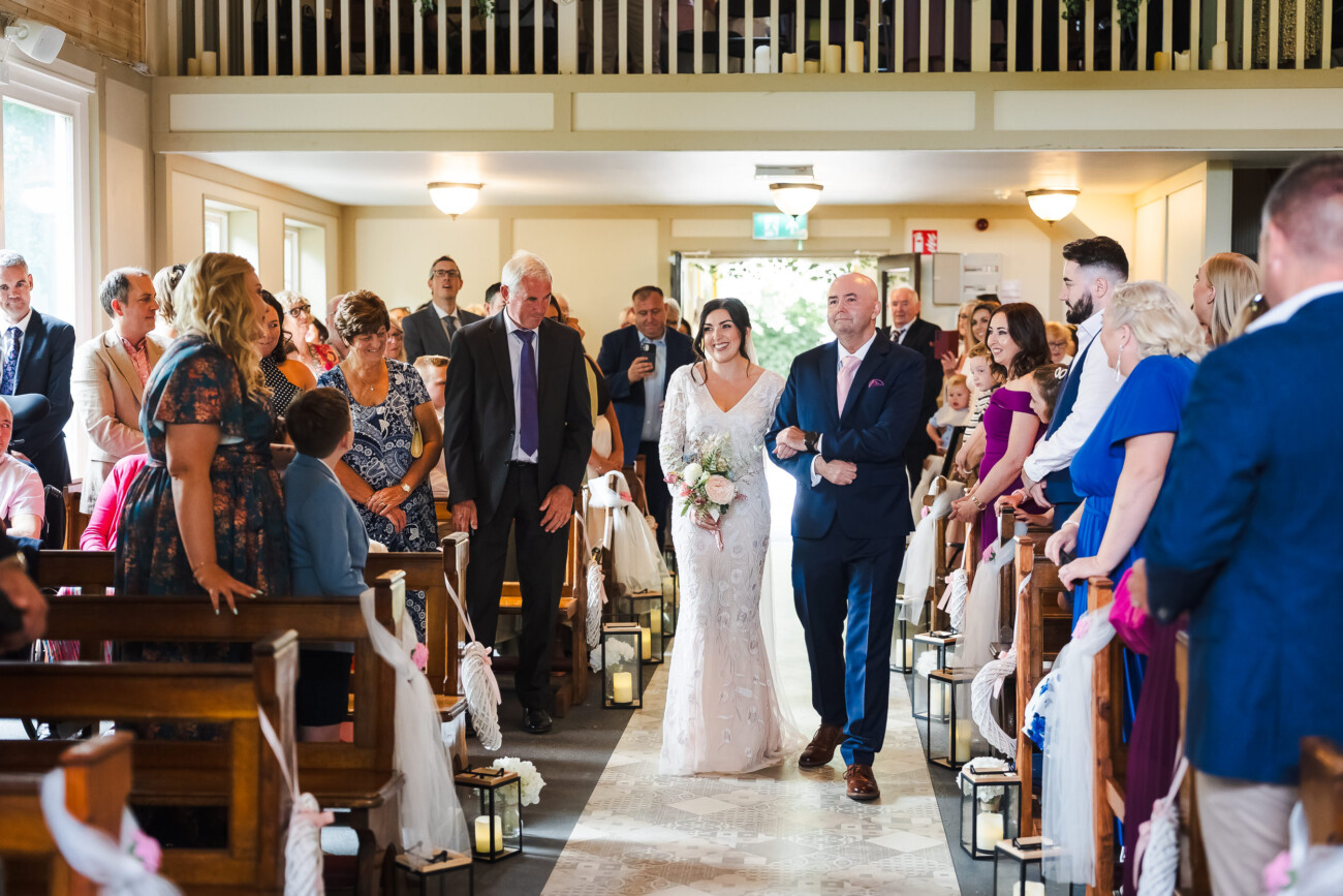 A Relaxed Summer Wedding at Village Barn, Tyrellspass | Amy & Eoghan 30 A bride in a white dress walks down the aisle with two men, surrounded by seated and standing wedding guests inside a decorated venue.