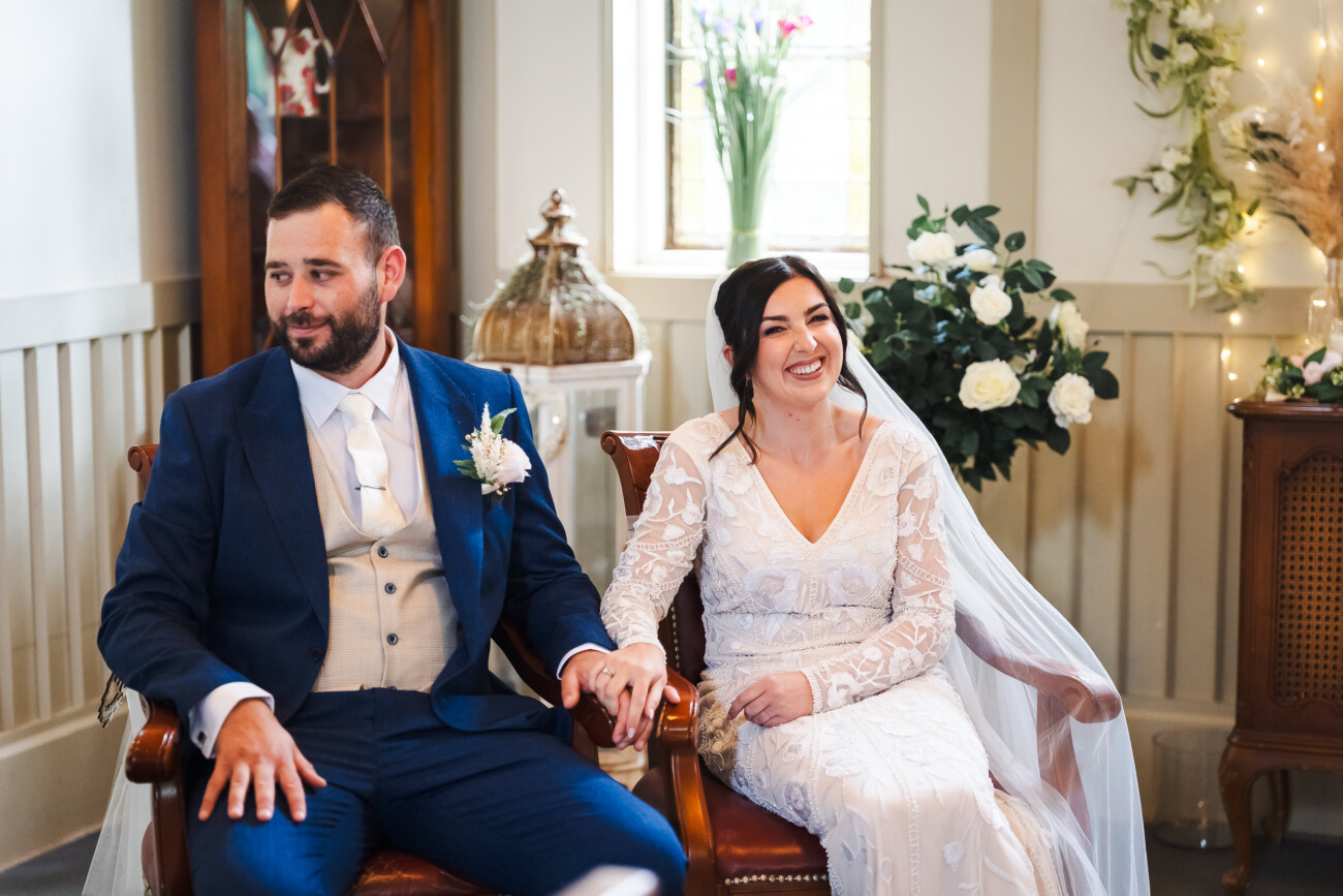 A Relaxed Summer Wedding at Village Barn, Tyrellspass | Amy & Eoghan 31 A bride and groom sit holding hands indoors, dressed in wedding attire, with flowers and decorations in the background.