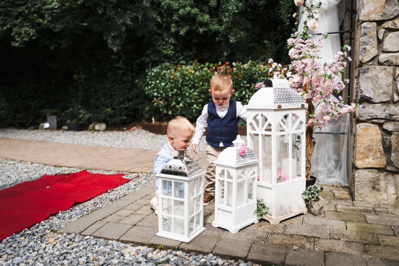 A Relaxed Summer Wedding at Village Barn, Tyrellspass | Amy & Eoghan 32 Two young boys in smart clothes kneel beside large white decorative lanterns and pink flowers on a paved area next to a stone wall and red carpet outdoors.