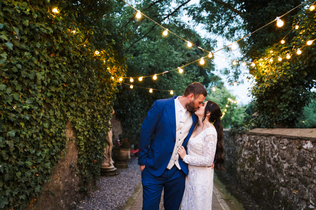 A Relaxed Summer Wedding at Village Barn, Tyrellspass | Amy & Eoghan 104 A couple in wedding attire kisses under fairy lights on a garden path, surrounded by ivy-covered walls and trees.