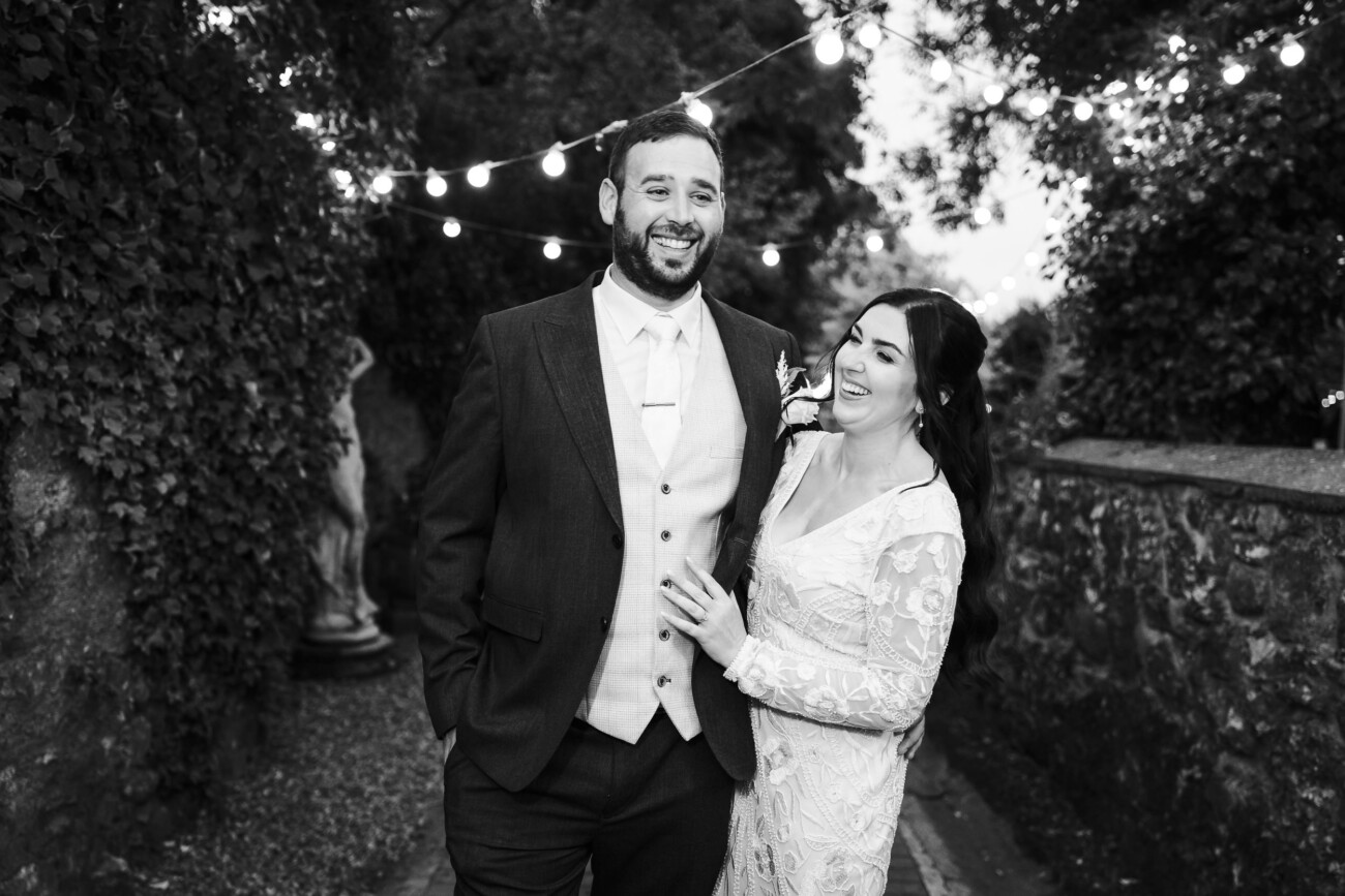 A Relaxed Summer Wedding at Village Barn, Tyrellspass | Amy & Eoghan 105 A man in a suit and a woman in a white dress stand outdoors, smiling under fairy lights, with trees and stone walls in the background.