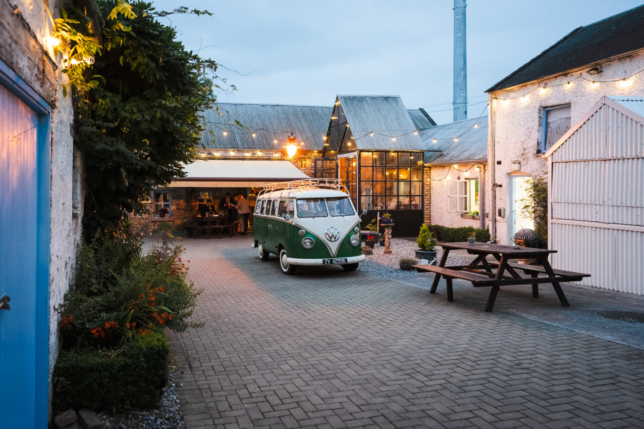 A Relaxed Summer Wedding at Village Barn, Tyrellspass | Amy & Eoghan 106 A vintage green and white Volkswagen van is parked in a courtyard surrounded by rustic buildings with fairy lights and outdoor seating.