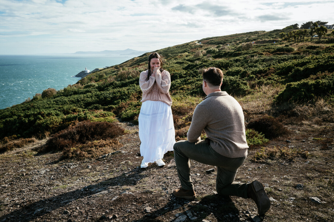 A Secret Proposal at Howth Cliffs & a Private Hour at Howth Castle: Joseph & Shannon's Engagement Shoot 2 A man kneels on one knee proposing to a surprised woman outdoors on a grassy hillside overlooking the sea.